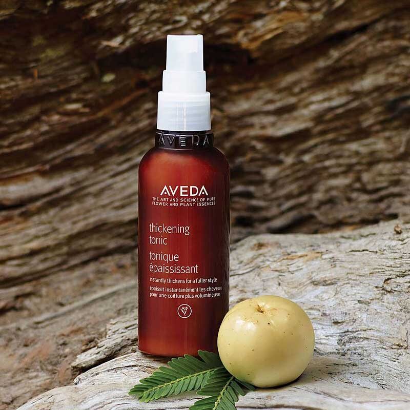 Aveda Thickening Tonic spray bottle on weathered wood beside a botanical fruit