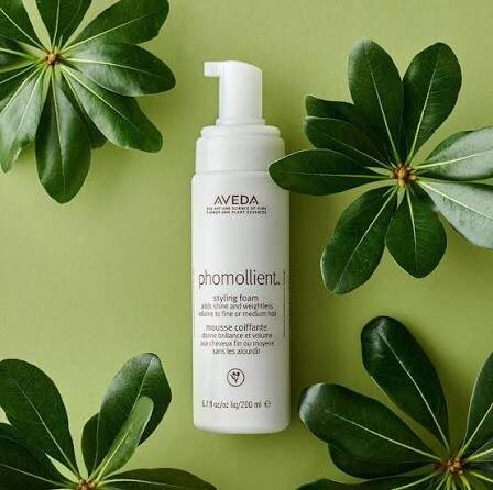 Aveda Phomollient Styling Foam bottle among glossy green leaves